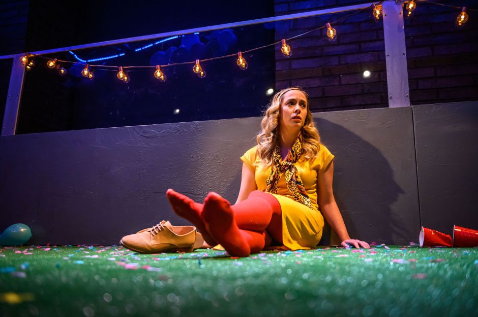 A woman looks upset as she sits on astroturf in a yellow dress and red tights